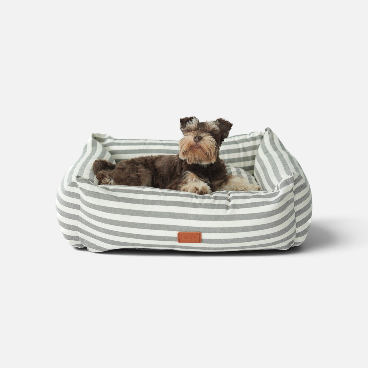 Cama para perros BIMBAS BRAND desenfundable con estampado de rayas grises y blancas, con un perro pequeño descansando.